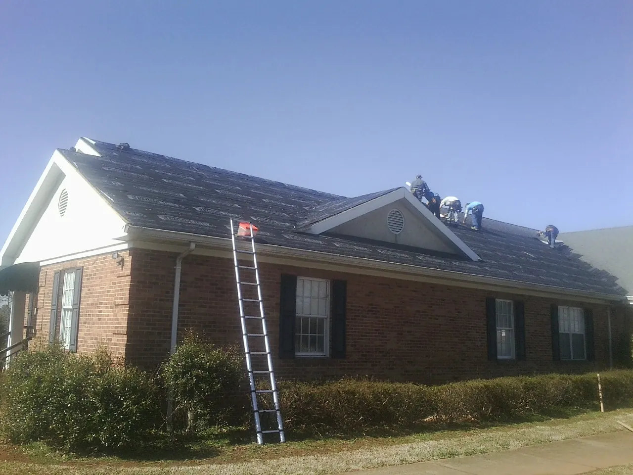 Crew tearing shingles off a brick ranch roof with a ladder set — Metal Roof Repair work by our crew in Goldens Bridge