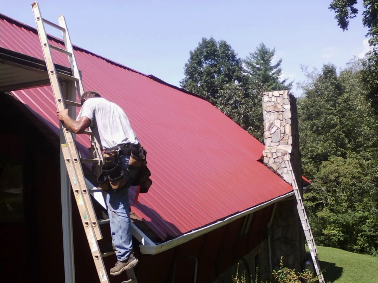 Roofer on a ladder beside a red metal roof and stone chimney — Soffit & Fascia Repair by Roofing Brothers in Goldens Bridge