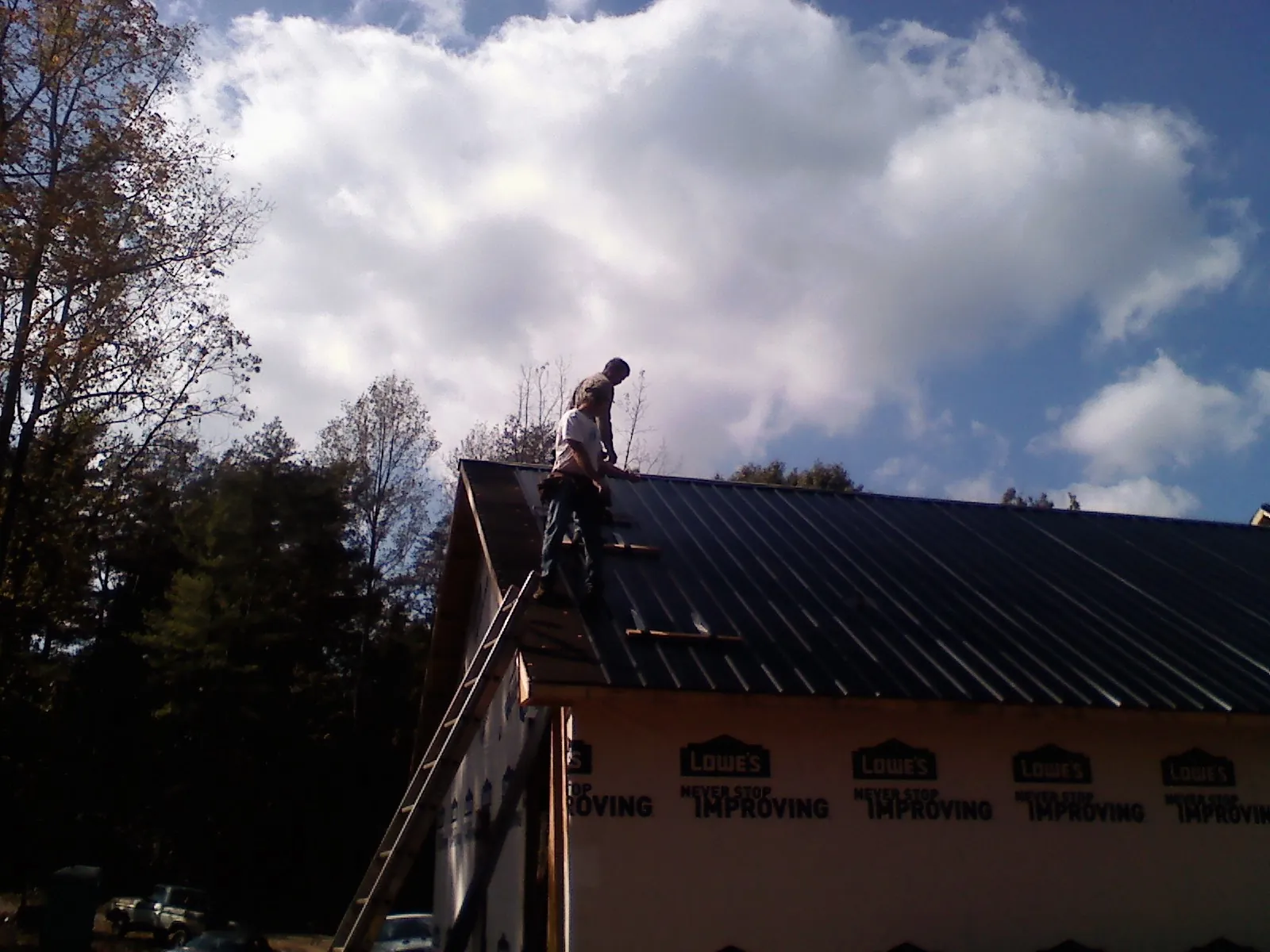 Roofer fastening dark metal panels high against a cloudy sky, from a Roof Inspection job in Goldens Bridge